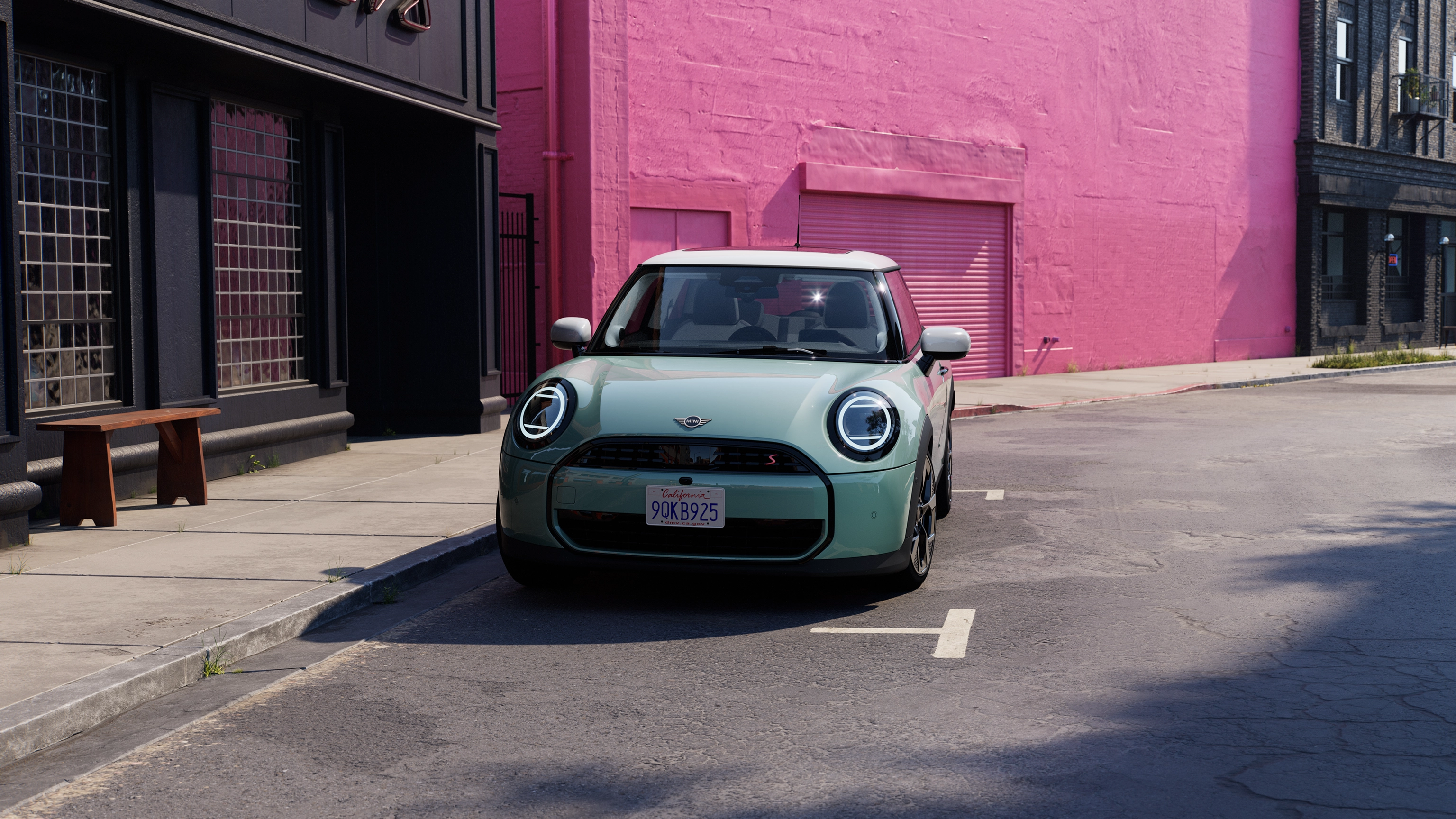Μπροστινή όψη MINI Cooper με χρώμα Ocean Wave Green και λευκή οροφή, το οποίο σταθμεύει σε δρόμο με ροζ τοίχο στο βάθος