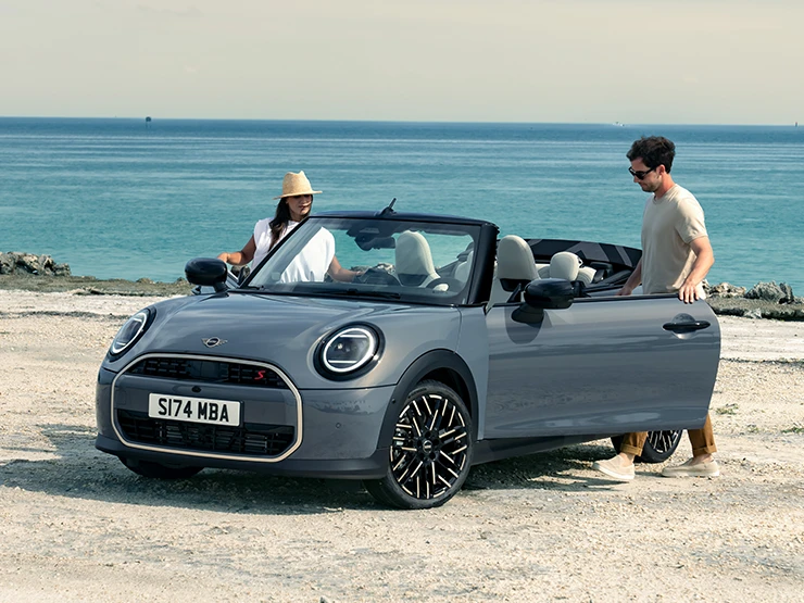 Μπροστινή αριστερή όψη του MINI Cooper Cabrio σε χρώμα Copper Grey σε παραλία με δύο άτομα δίπλα του.