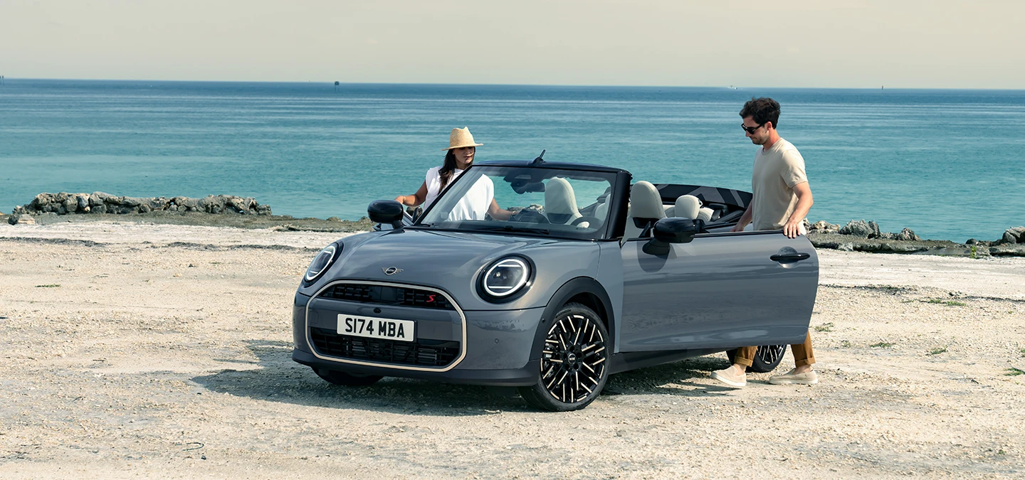 Μπροστινή αριστερή όψη του MINI Cooper Cabrio σε χρώμα Copper Grey σε παραλία με δύο άτομα δίπλα του.