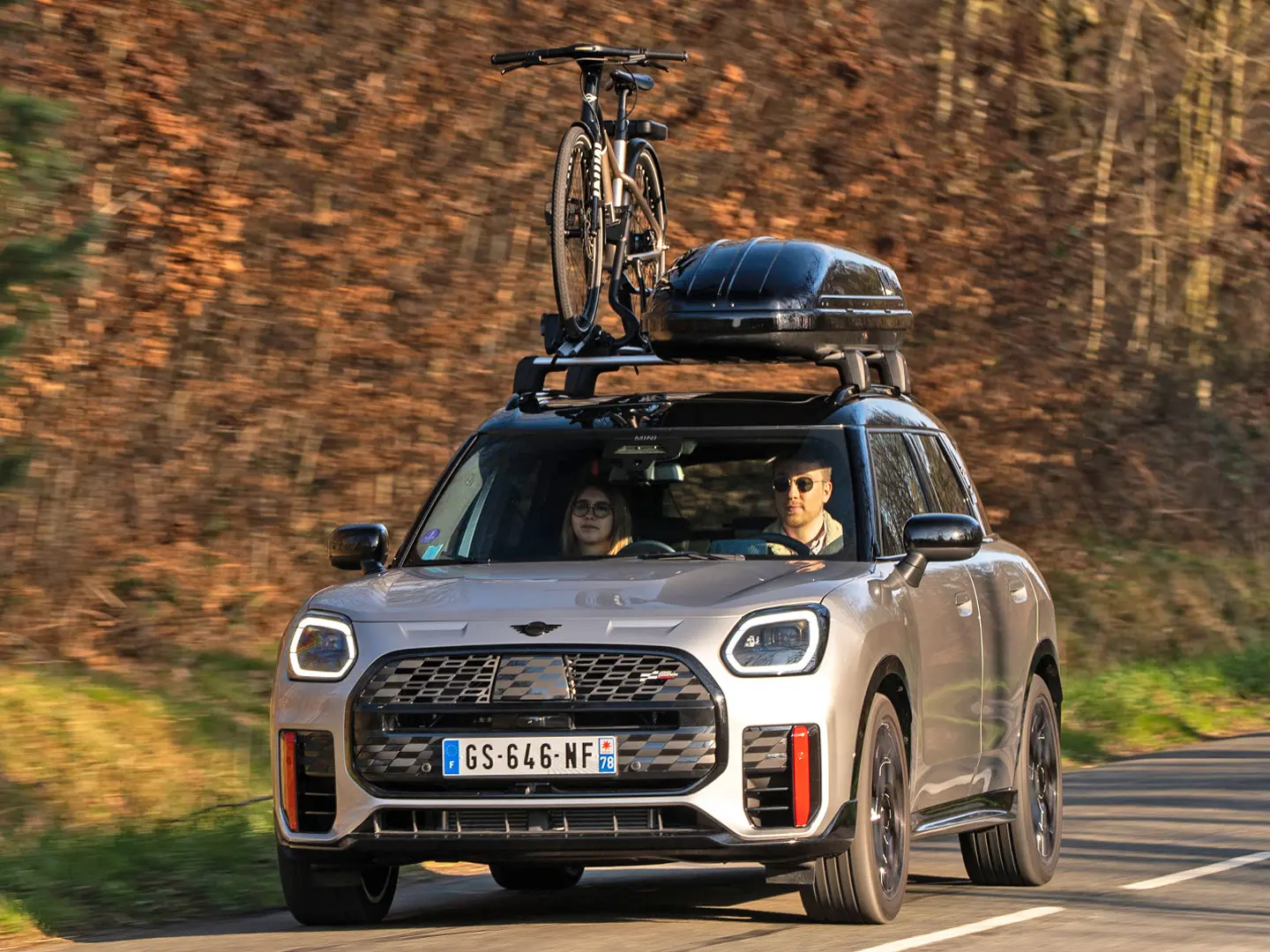 MINI Countryman Cooper σε Silver σε ορεινό δρόμο με φορέα οροφής MINI, μπαγκαζιέρα οροφής MINI, βάση μεταφοράς ποδηλάτων MINI με τοποθετημένο ποδήλατο και αεροτομή MINI John Cooper Works – μπροστινή όψη.