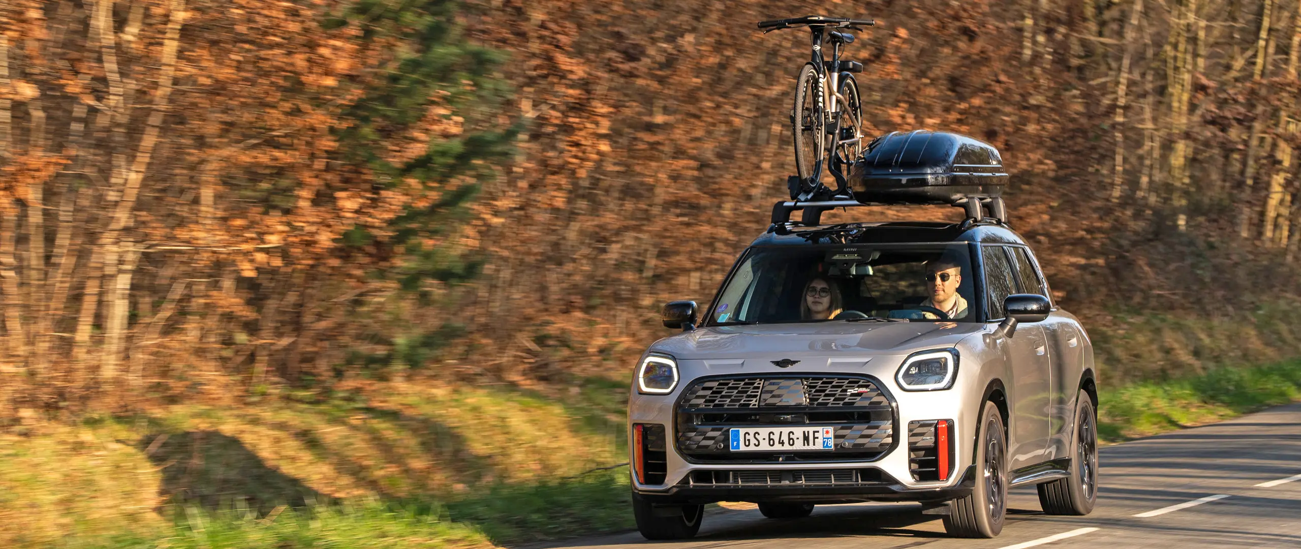MINI Countryman Cooper σε Silver σε ορεινό δρόμο με φορέα οροφής MINI, μπαγκαζιέρα οροφής MINI, βάση μεταφοράς ποδηλάτων MINI με τοποθετημένο ποδήλατο και αεροτομή MINI John Cooper Works – μπροστινή όψη.