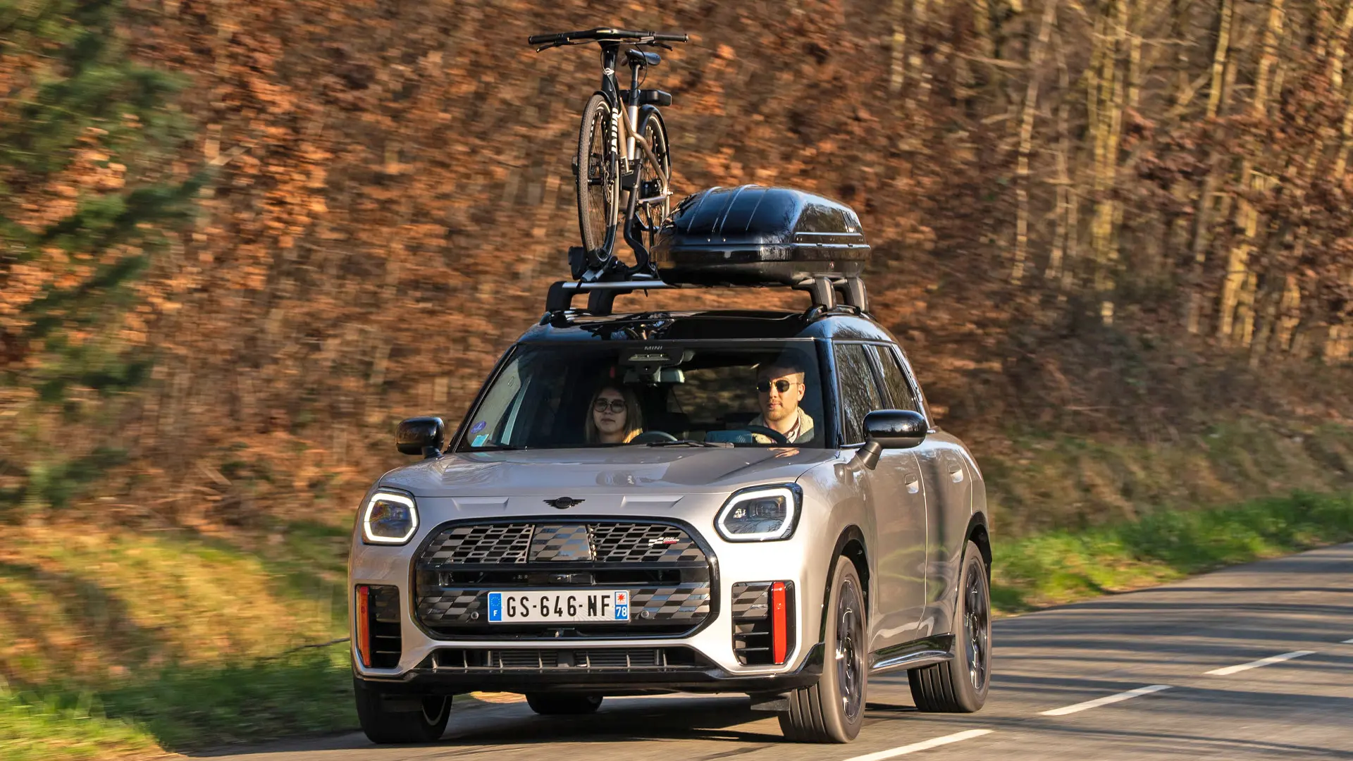 MINI Countryman Cooper σε Silver σε ορεινό δρόμο με φορέα οροφής MINI, μπαγκαζιέρα οροφής MINI, βάση μεταφοράς ποδηλάτων MINI με τοποθετημένο ποδήλατο και αεροτομή MINI John Cooper Works – μπροστινή όψη.