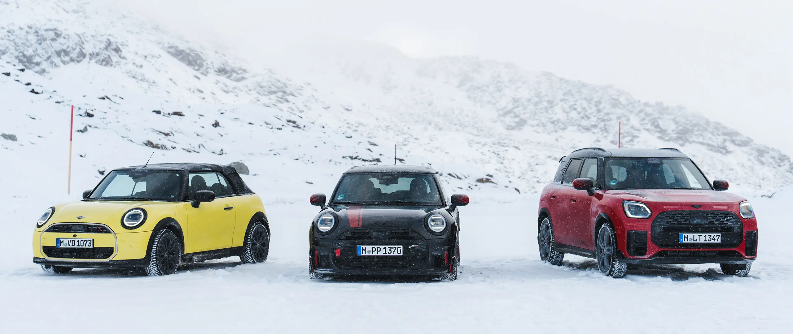 MINI Cooper Cabrio σε Sunny Side Yellow, MINI John Cooper Works σε Midnight Black II και MINI Countryman Cooper σε Chilli Red – σταθμευμένα σε χιονισμένο ορεινό δρόμο, μπροστινή όψη.