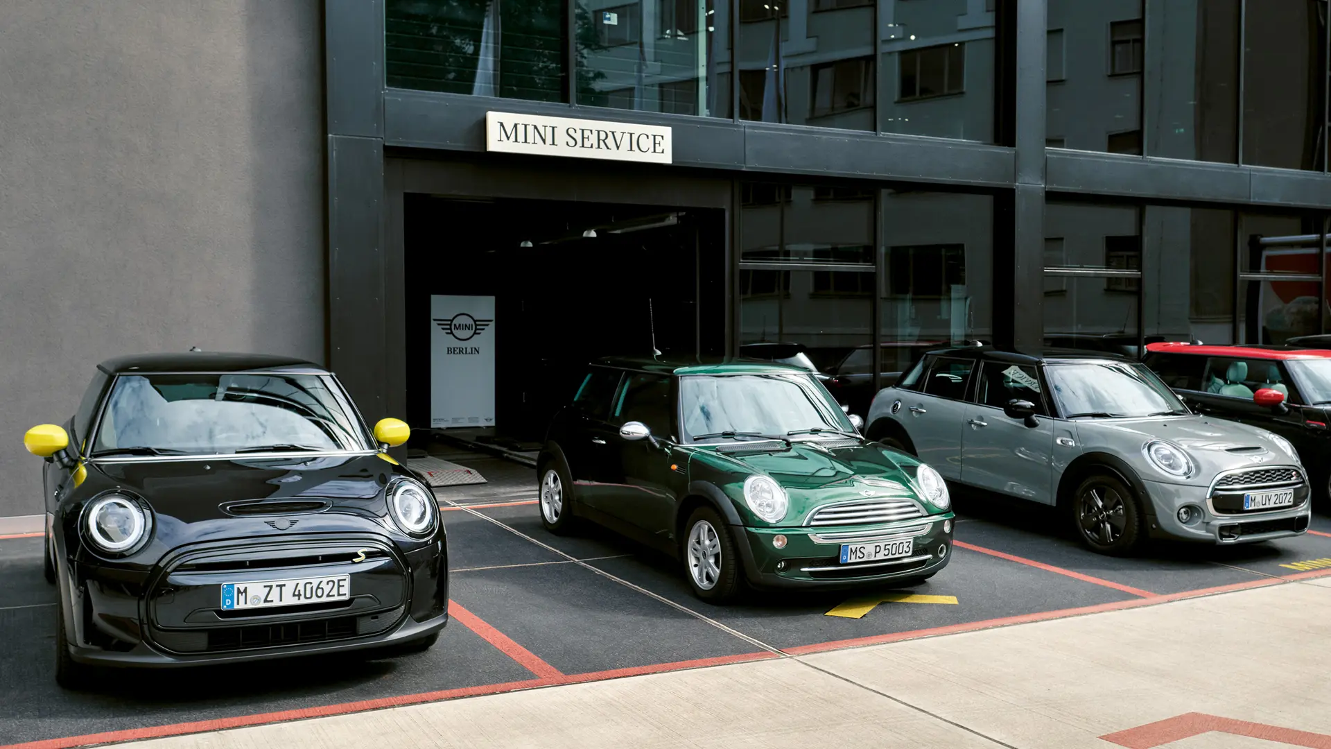 Ηλεκτρικό MINI Cooper S σε μαύρο χρώμα, σε εξωτερικό χώρο Επίσημου Εμπόρου MINI, μαζί με τρίθυρο MINI Cooper σε πράσινο χρώμα, πεντάθυρο MINI σε ασημί χρώμα, MINI JCW σε μαύρο χρώμα, μπροστινή όψη. 