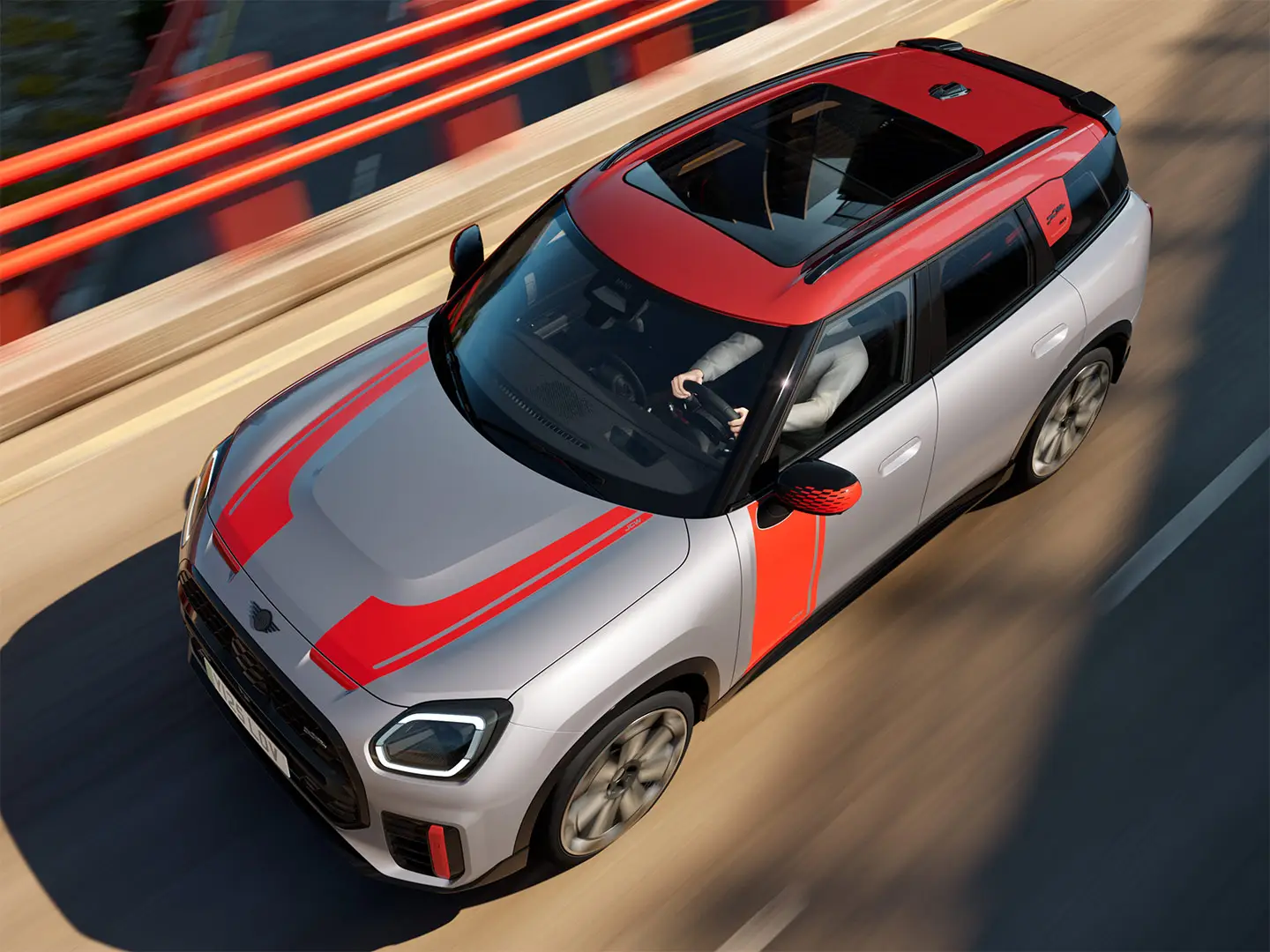 Ένα MINI Countryman John Cooper Works σε Melting Silver με καλύμματα εξωτερικών καθρεπτών, πλευρικά αυτοκόλλητα και αυτοκόλλητο στο καπό του κινητήρα, αεροτομή οροφής και πτερύγια αεροτομής σε Chili Red κινείται σε έναν αστικό δρόμο – μπροστινή όψη από το πλάι.