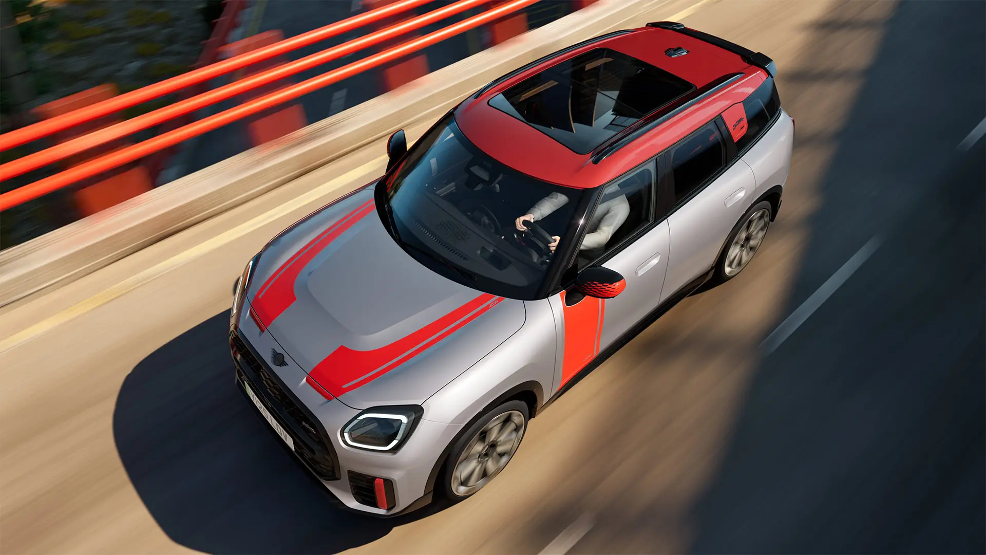Ένα MINI Countryman John Cooper Works σε Melting Silver με καλύμματα εξωτερικών καθρεπτών, πλευρικά αυτοκόλλητα και αυτοκόλλητο στο καπό του κινητήρα, αεροτομή οροφής και πτερύγια αεροτομής σε Chili Red κινείται σε έναν αστικό δρόμο – μπροστινή όψη από το πλάι.