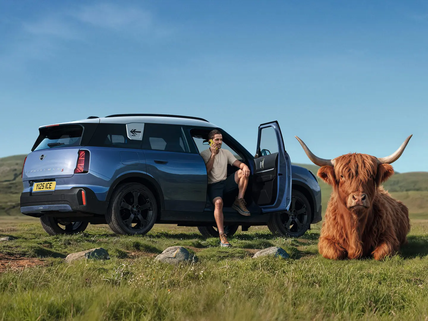 Ένας άνδρας έξω από το MINI Countryman S του μιλά με τη MINI Roadside Assistance μέσω της εφαρμογής MINI App και ένας ταύρος των Χάιλαντς είναι ξαπλωμένος εκεί κοντά.